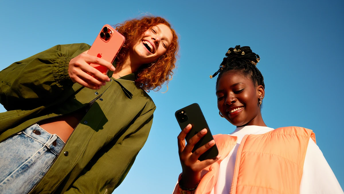 Zwei Freundinnen lachen mit ihren Smartphones unter blauem Himmel.